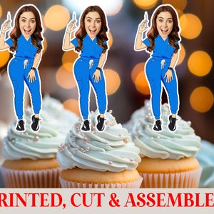 May include: Three cupcakes topped with cartoon figures of a woman in blue scrubs holding a syringe. The cupcakes have light blue frosting and are decorated with small white pearls. The text "PRINTED, CUT & ASSEMBLED" is at the bottom.