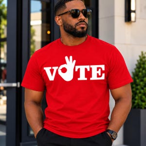 以下が含まれることがあります： 胸元に大きな白い大文字でVOTEとプリントされた鮮やかな赤の半袖Tシャツ。Oの文字は、OKサインをしている手の白いグラフィックに置き換えられています。このTシャツはクルーネックで、標準的なフィット感のデザインです。