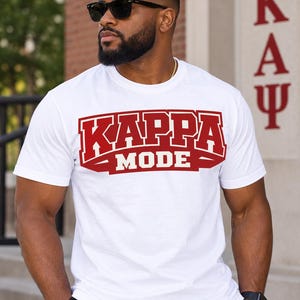 Può includere: Una t-shirt bianca a maniche corte con girocollo e una grande grafica rosso scuro sul petto. La grafica mostra la parola KAPPA in un carattere in stile college sopra la parola MODE scritta in caratteri bianchi più piccoli.