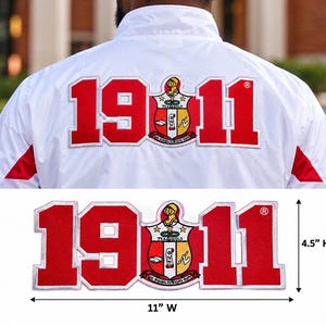 May include: A white jacket featuring a large red embroidered patch on the back with the text 1911 and a central crest. The patch measures 11 inches wide by 4.5 inches high, as indicated by the visible measurement markings.