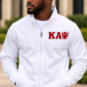 May include: A white long-sleeve zip-up jacket featuring a high collar and two front pockets. On the left chest, there is a red embroidered patch with white trim displaying the Greek letters Kappa Alpha Psi, written as KAY.