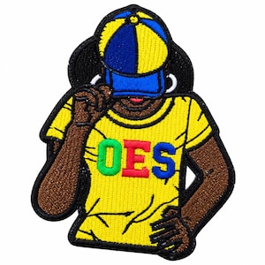 May include: An embroidered patch featuring a figure wearing a yellow and blue baseball cap and a yellow t-shirt with the letters OES in green, red, and blue. The figure has brown skin, hoop earrings, and is depicted from the chest up in a pose.