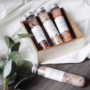 May include: Four small glass tubes filled with colorful bath salts, labeled "Sleepy Beauty", "Cranberry & Ginger", "Blackberry", and "Sea Salt". The tubes are in a wooden box on a white fabric surface.