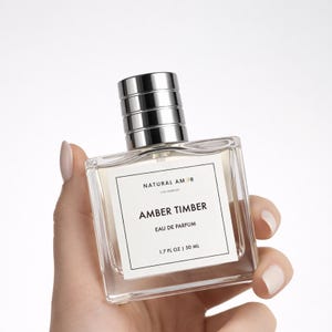 May include: A clear glass perfume bottle with a silver cap and a white label that reads "NATURAL AMOR LOS ANGELES AMBER TIMBER EAU DE PARFUM 1.7 FL OZ | 50 ML". The bottle is held by a hand with manicured nails.