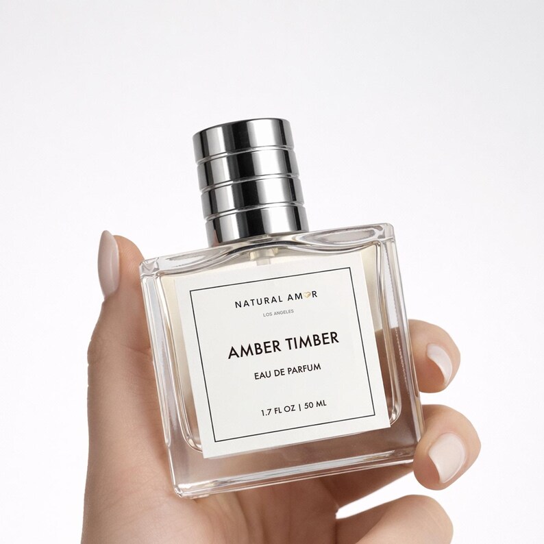 May include: A clear glass perfume bottle with a silver cap and a white label that reads "NATURAL AMOR LOS ANGELES AMBER TIMBER EAU DE PARFUM 1.7 FL OZ | 50 ML". The bottle is held by a hand with manicured nails.