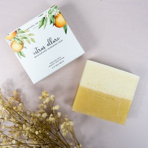May include: A white box with a citrus illustration and the text "NATURAL AMOR" and "citrus allure MOISTURIZING HANDMADE SOAP ENRICHED WITH ORGANIC SHEA BUTTER 3.5 OZ/100 G". A white and yellow bar of soap with the text "NATURAL AMOR" on it.