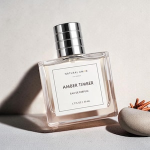 May include: A clear, rectangular perfume bottle with a silver cap. The label reads "NATURAL AMOR" and "AMBER TIMBER EAU DE PARFUM 1.7 FL OZ | 50 ML." A small stone and dried plant are next to the bottle. The perfume bottle is angled.