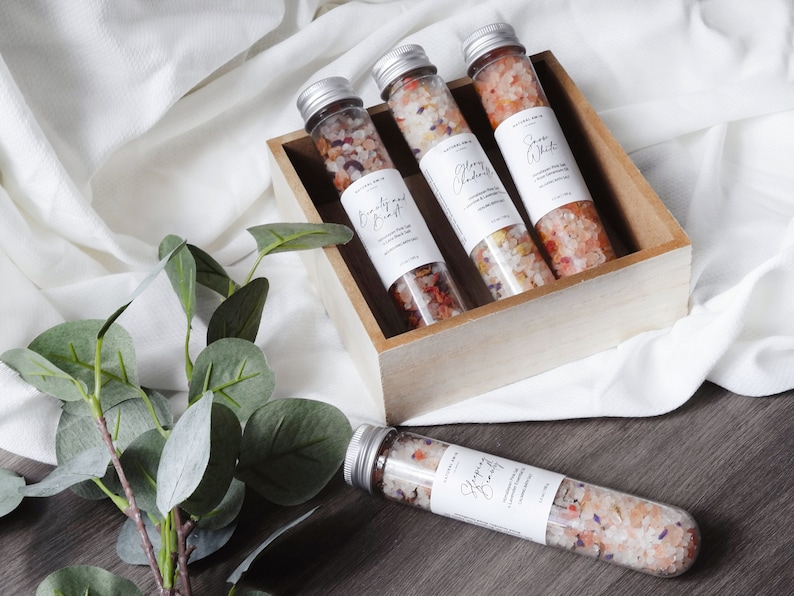 May include: Four small glass tubes filled with colorful bath salts, labeled "Sleepy Beauty", "Cranberry & Ginger", "Blackberry", and "Sea Salt". The tubes are in a wooden box on a white fabric surface.