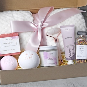 May include: A gift box containing a white textured blanket tied with a pink ribbon, a Rose Honey soap bar, two bath bombs, a Rose Bouquet soy candle, a rose quartz facial roller, a tube of lavender hand cream, and a jar of dried floral facial steam.