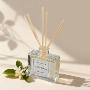 May include: A clear glass reed diffuser with wooden reeds. The label reads "NATURAL AMOR COCONUT CREAM REED DIFFUSER". A sprig of white flowers and green leaves is in the lower left corner. The background is a neutral beige.
