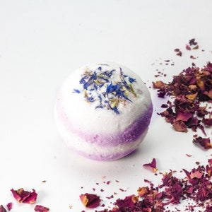 Pode incluir: Uma bomba de banho branca com um redemoinho roxo e p&eacute;talas de flores azuis e verdes no topo. A bomba de banho est&aacute; rodeada de p&eacute;talos de rosa secos.