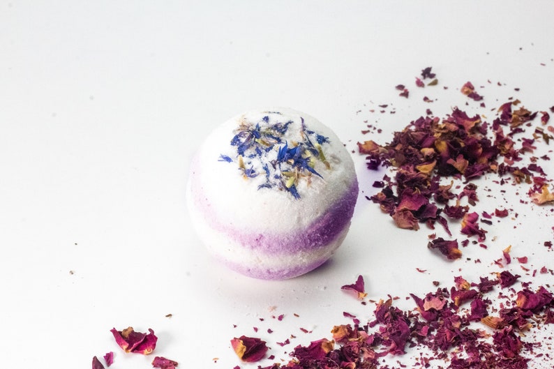 Pode incluir: Uma bomba de banho branca com um redemoinho roxo e p&eacute;talas de flores azuis e verdes no topo. A bomba de banho est&aacute; rodeada de p&eacute;talos de rosa secos.