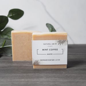 May include: Two bars of handmade soap with a light brown color. The soap bar on top has a white label with the text "NATURAL AMOR LOS ANGELES MINT COFFEE AMOR HANDMADE SOAP BAR | 4.3 OZ"