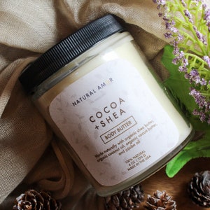 May include: A jar of cocoa and shea body butter with a black lid and a white label. The label reads "NATURAL AMOR COCOA + SHEA BODY BUTTER." The jar is 6 oz / 170 g. The jar is on a beige cloth with purple flowers.