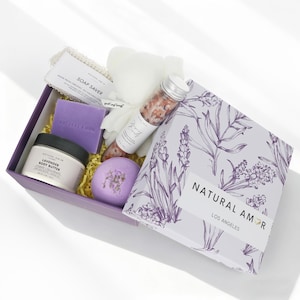 May include: A lavender-themed gift set in a purple box. The box contains a jar of body butter, a bar of soap, a bath bomb, a tube of bath salts, a soap saver, and a loofah. The box is decorated with lavender sprigs and the text "NATURAL AMOR LOS ANGELES".