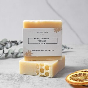 May include: Two bars of handmade soap stacked on top of each other. The top bar has a white label with the text "Natural Am-Jr" and "Honey Orange Turmeric - Amor" and "Handmade Soap Bar | 4.3 oz". The bottom bar is a light yellow color and has a honeycomb pattern.