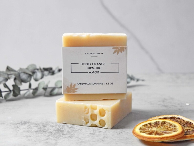 May include: Two bars of handmade soap stacked on top of each other. The top bar has a white label with the text "Natural Am-Jr" and "Honey Orange Turmeric - Amor" and "Handmade Soap Bar | 4.3 oz". The bottom bar is a light yellow color and has a honeycomb pattern.