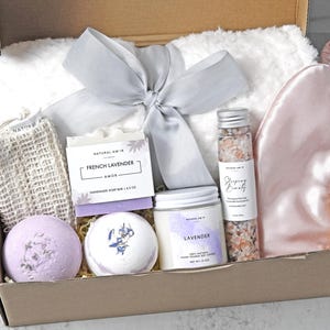 May include: A gift box containing a white faux fur blanket with a grey ribbon, a pink satin sleep mask, a beige mesh soap pouch, a French Lavender soap bar, two round bath bombs, a jar of lavender soy candle, and a tube of pink bath salts.