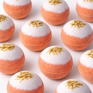 May include: A collection of spherical bath bombs. Each bath bomb is a combination of white, orange, and gold. The top half is white, the bottom half is orange, and the top is decorated with gold flakes. The bath bombs are arranged in a grid pattern.