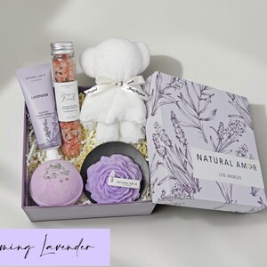 Puede incluir: Un set de regalo que contiene un tubo de crema de manos de lavanda, un tubo de cristal con sales de ba&ntilde;o rosas, un oso de peluche blanco, una bomba de ba&ntilde;o morada redonda y una vela con forma de flor morada en un plato negro, dentro de una caja.
