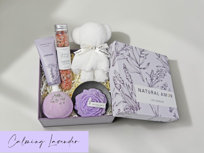 Puede incluir: Un set de regalo que contiene un tubo de crema de manos de lavanda, un tubo de cristal con sales de ba&ntilde;o rosas, un oso de peluche blanco, una bomba de ba&ntilde;o morada redonda y una vela con forma de flor morada en un plato negro, dentro de una caja.