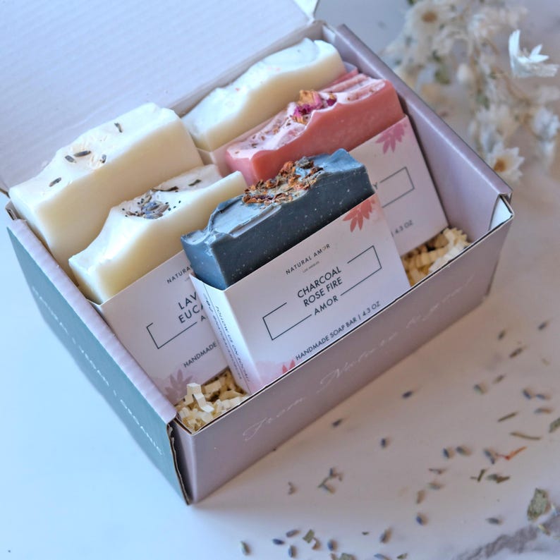 May include: A gift box filled with four bars of handmade soap. The soaps are labeled "Lavender Eucalyptus", "Charcoal Rose Fire Amor", and two other unreadable labels. The box is filled with shredded paper and has a pink and green color scheme.
