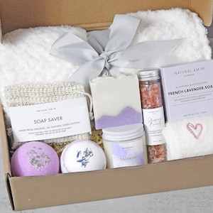 May include: A gift box filled with self-care items. Includes a white fluffy towel, a bar of lavender soap, a jar of bath salts, a candle, a soap saver, and two bath bombs. The box is tied with a gray ribbon.