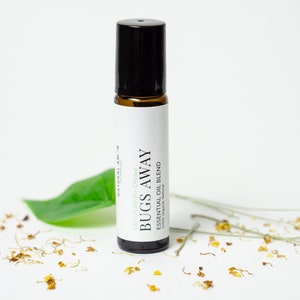May include: A small glass bottle with a black rollerball top. The bottle is labeled "BUGS AWAY 100% Pure + Dried ESSENTIAL OIL BLEND". The bottle is sitting on a white surface with green leaves and yellow flower petals.
