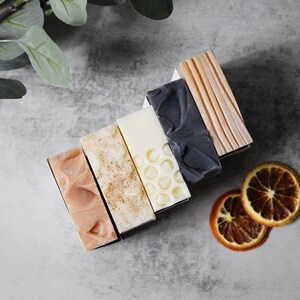 May include: Six bars of handmade soap arranged in a stack. The bars are in various colors and patterns, including pink, brown, white, and black. The soap bars are on a gray surface with dried orange slices.