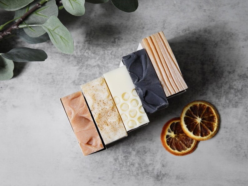 May include: Six bars of handmade soap arranged in a stack. The bars are in various colors and patterns, including pink, brown, white, and black. The soap bars are on a gray surface with dried orange slices.
