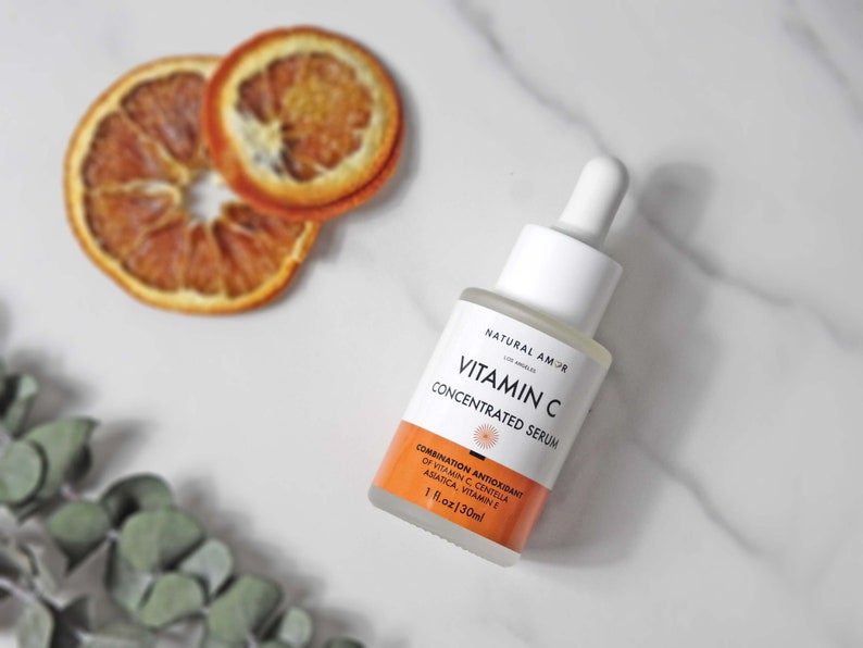 May include: A white and clear glass bottle of Vitamin C Concentrated Serum with a white dropper. The bottle has orange and white labels. Two dried orange slices are in the background. The bottle contains 1 fl oz or 30ml.