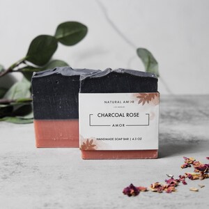 May include: Two bars of handmade soap, one black and one pink, with a label that reads "Natural Amor, Los Angeles, Charcoal Rose, Amor, Handmade Soap Bar | 4.3 oz."