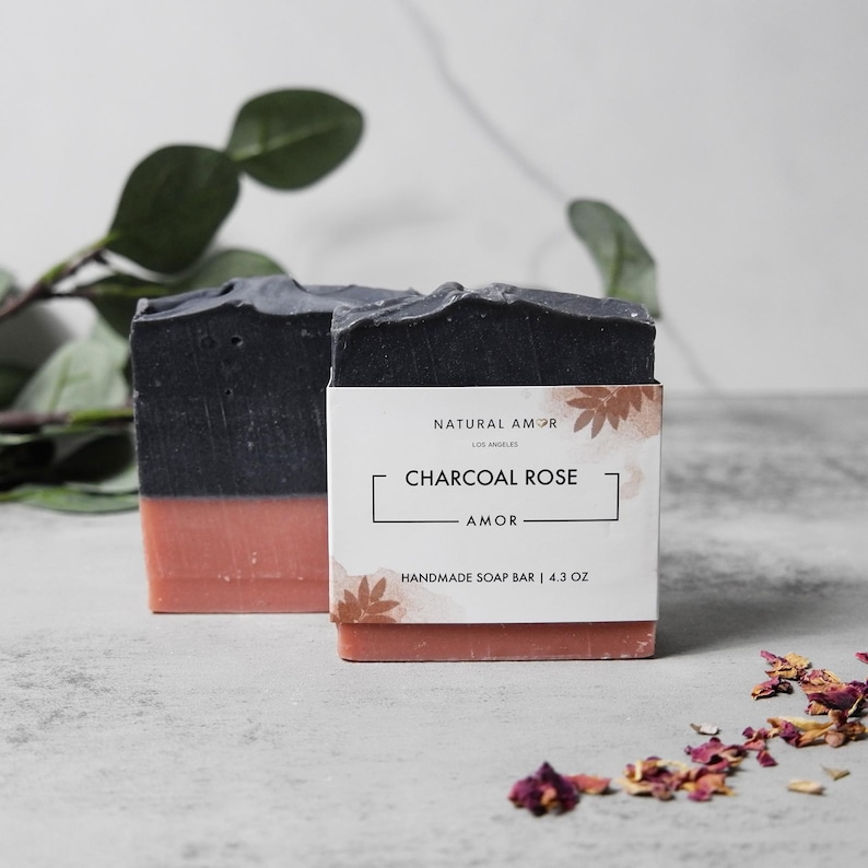 May include: Two bars of handmade soap, one black and one pink, with a label that reads "Natural Amor, Los Angeles, Charcoal Rose, Amor, Handmade Soap Bar | 4.3 oz."