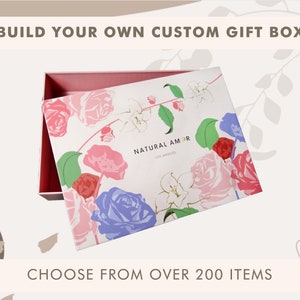 May include: A white gift box with a floral design featuring pink, red, blue, and green roses. The box is open and the lid is propped up. The text "NATURAL AMOR" and "LOS ANGELES" is printed on the lid. The text "BUILD YOUR OWN CUSTOM GIFT BOX" is printed above the box and the text "CHOOSE FROM OVER 200 ITEMS" is printed below the box.