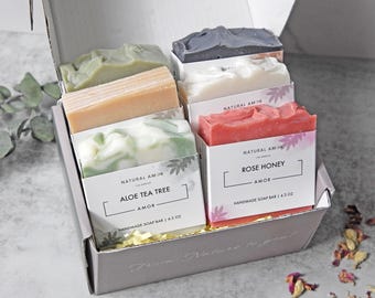 May include: A gift box filled with handmade soap bars. The box contains bars in various colors, including green, beige, white, and red. Each bar is labeled with the scent and brand name. The box is lined with yellow crinkle paper.