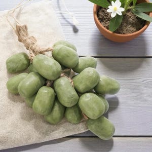 May include: A bunch of green oval-shaped soaps tied together with twine, resembling grapes, rests on a beige cloth. A small potted plant with a white flower is in the background. The soaps have a smooth, matte finish.