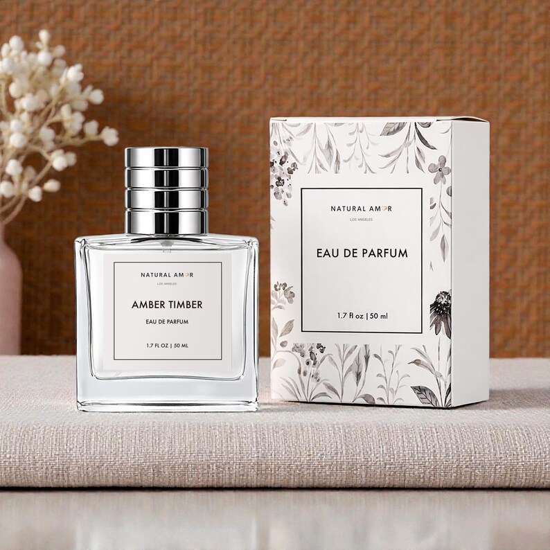 May include: A clear glass perfume bottle with a silver cap and a matching box. The bottle label reads "AMBER TIMBER EAU DE PARFUM 1.7 fl oz | 50 ml." The box is white with a floral design and the text "NATURAL AMOR EAU DE PARFUM 1.7 fl oz | 50 ml."