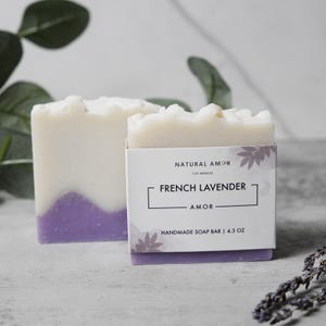 May include: Two bars of handmade soap, one with a purple swirl and the other with a white label that reads "Natural Amor Los Angeles French Lavender Amor Handmade Soap Bar | 4.3 oz"