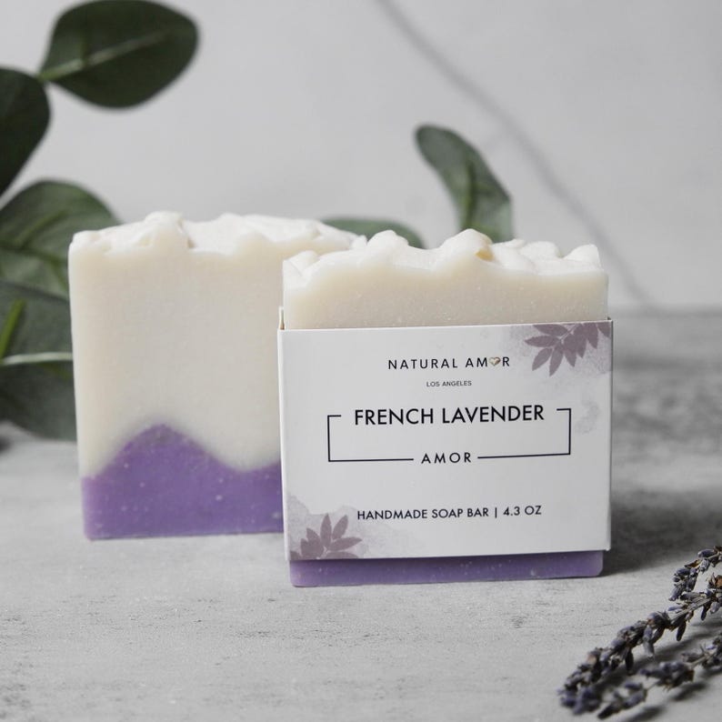 May include: Two bars of handmade soap, one with a purple swirl and the other with a white label that reads "Natural Amor Los Angeles French Lavender Amor Handmade Soap Bar | 4.3 oz"