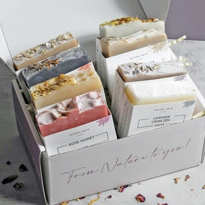 May include: A white gift box filled with various handmade soaps. The soaps are in different colors and textures, some with visible botanicals. The box has the text "From Nature to you!" and "Rose Honey" and "Lavender Cedar Zen" on the labels.