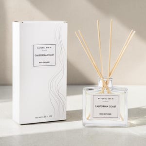 May include: A white box and a clear glass reed diffuser. The box has the text "NATURAL AM CALIFORNIA COAST REED DIFFUSER" and a decorative wave design. The diffuser has wooden reeds and the same text on a label. The bottle contains 100 ml / 3.34 fl oz.