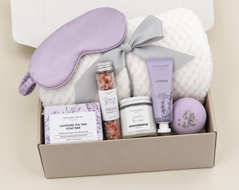 May include: A gift box containing a lavender sleep mask, a white textured blanket with a grey ribbon, a bar of lavender tea tree soap, a glass vial of pink bath salts, a small candle, a tube of lavender hand cream, and a round lavender bath bomb.