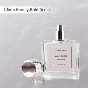 May include: A clear glass perfume bottle with a silver cap and sprayer, labeled "AMBER TIMBER" by "NATURAL AMOR." The bottle contains a light-colored liquid and is 1.7 fl oz / 50 ml. The text "Clean Beauty, Bold Scent" is at the top.