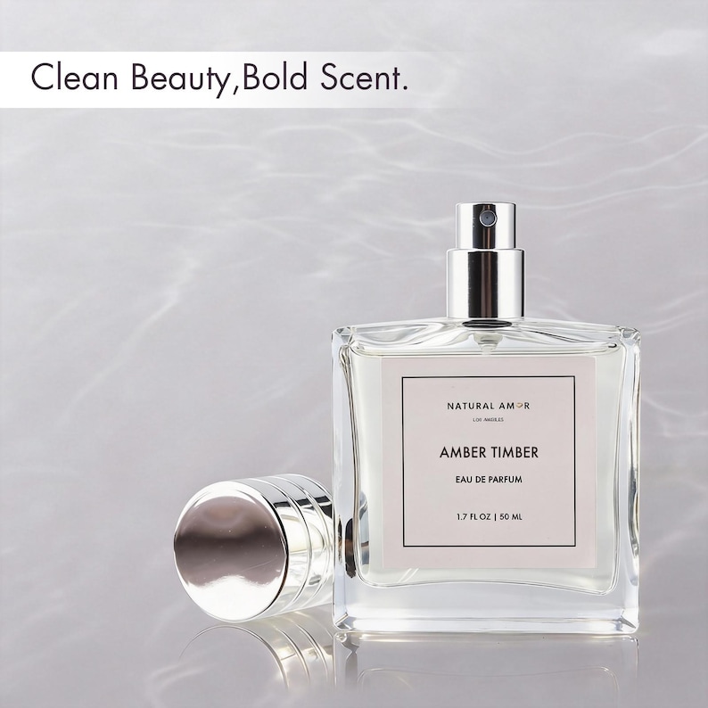 May include: A clear glass perfume bottle with a silver cap and sprayer, labeled "AMBER TIMBER" by "NATURAL AMOR." The bottle contains a light-colored liquid and is 1.7 fl oz / 50 ml. The text "Clean Beauty, Bold Scent" is at the top.