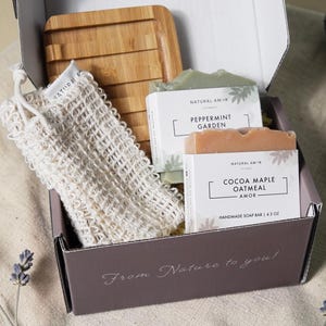 May include: A gift box containing handmade soap bars, a wooden soap dish, and a natural fiber washcloth. The soap bars are labeled "Peppermint Garden" and "Cocoa Maple Oatmeal". The box is printed with "From Nature to you!"