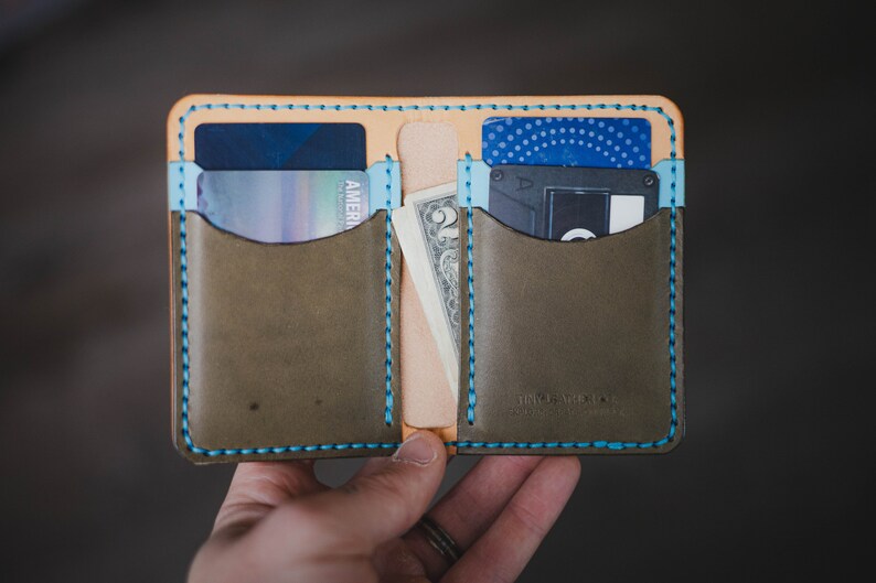 Op de afbeelding: Een bruine leren portemonnee met blauwe stiksels en twee kaartsleuven. De portemonnee is open en bevat een creditcard, contant geld en een kleine zwarte kaarthouder.