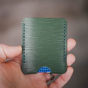 May include: A green leather card holder with a blue and white patterned card inside. The card holder has a stitched edge.
