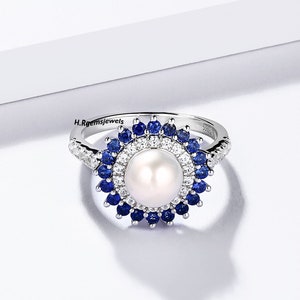 May include: A silver ring featuring a large white pearl encircled by a halo of smaller clear and blue gemstones.