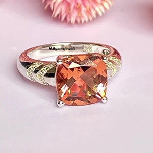 Peut inclure: Une bague en argent avec une grosse pierre précieuse carrée de couleur pêche. La bande présente un motif texturé avec de petits accents scintillants. La bague est posée sur un fond rose clair, avec le texte "H.Rgemsjewels" visible.