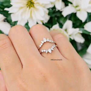 May include: A silver ring guard with a curved design, set with multiple small, clear gemstones. The ring guard is designed to be worn with a ring. The background features white flowers and green foliage.